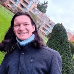 Paul outside in a coat, grinning playfully, the houses in the background and the trees except one next to Paul are at a 45° angle